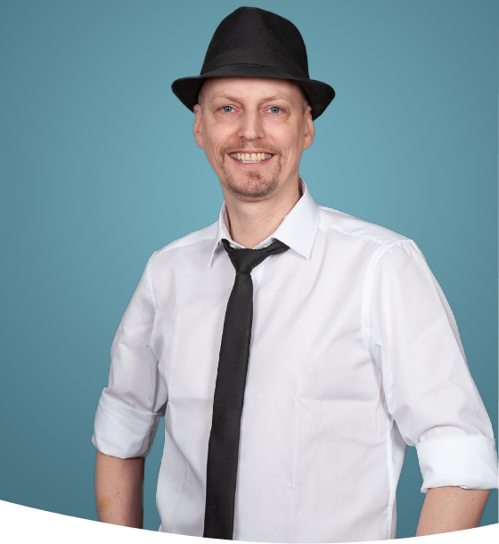 Mann in weißem Hemd und schwarzer Krawatte mit Fedora-Hut lächelt vor blauem Hintergrund.