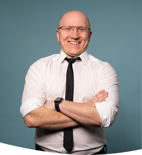 Glatzköpfiger Mann mit lächelnder Brille, weißem Hemd und schwarzer Krawatte, der mit verschränkten Armen vor einem blaugrünen Hintergrund steht.