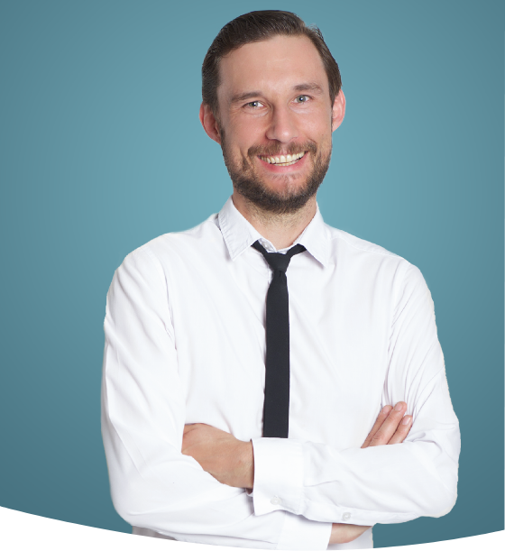 Ein lächelnder Mann in weißem Hemd und schwarzer Krawatte mit verschränkten Armen vor blauem Hintergrund.
