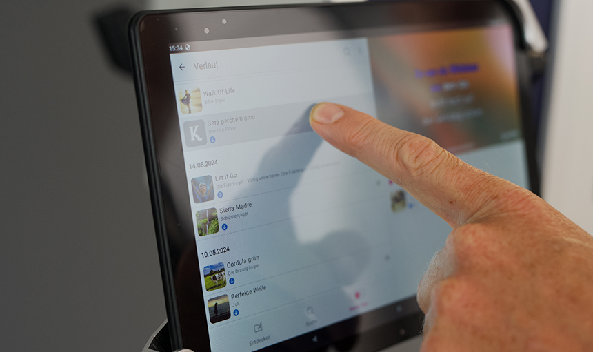 Eine Person benutzt ein Tablet und scrollt durch eine Playlist mit Liedern. Der Finger zeigt auf ein Lied namens „Skal prøvés å amo“ von Les Enfoirés. Eine Person benutzt ein Tablet und scrollt durch eine Playlist mit Liedern. Der Finger zeigt auf ein Lied namens „Skal prøvés å amo“ von Les Enfoirés.