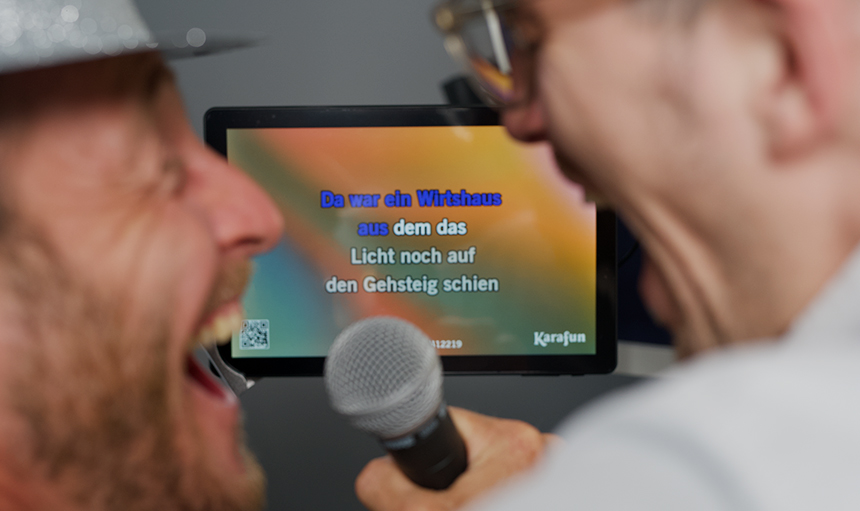 Zwei Personen singen in ein Mikrofon, im Hintergrund läuft auf einem Karaoke-Bildschirm ein deutscher Liedtext. Zwei Personen singen in ein Mikrofon, im Hintergrund läuft auf einem Karaoke-Bildschirm ein deutscher Liedtext.