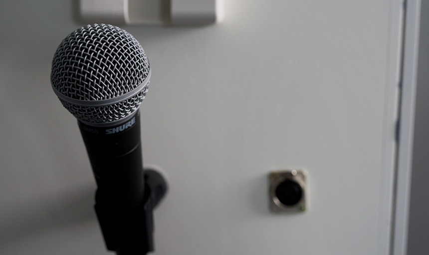 Ein Mikrofon auf einem Ständer steht vor einer weißen Wand, im Hintergrund ist eine kleine quadratische Wandsteckdose zu sehen. Ein Mikrofon auf einem Ständer steht vor einer weißen Wand, im Hintergrund ist eine kleine quadratische Wandsteckdose zu sehen.