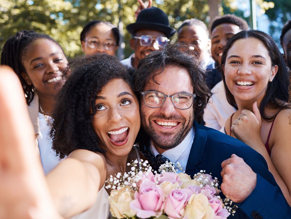 Ein frisch vermähltes Paar macht draußen ein Selfie mit lächelnden Freunden. Die Braut hält einen Strauß rosa und gelber Rosen in der Hand. Alle sehen glücklich und feierlich aus. Ein frisch vermähltes Paar macht draußen ein Selfie mit lächelnden Freunden. Die Braut hält einen Strauß rosa und gelber Rosen in der Hand. Alle sehen glücklich und feierlich aus.