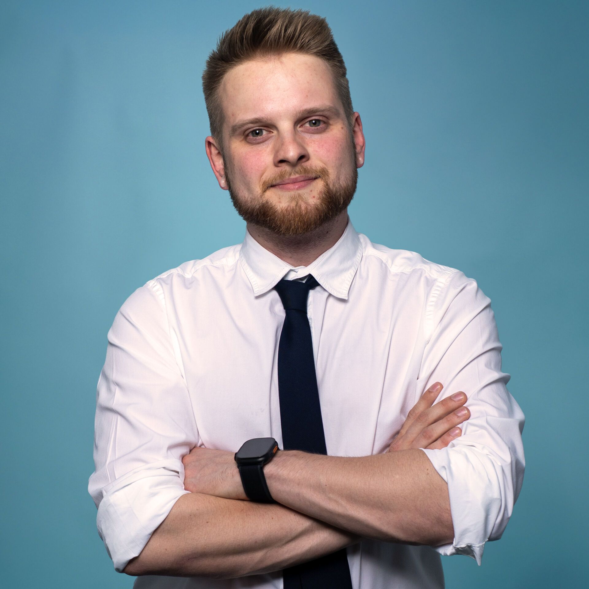 Ein Mann mit hellem Haar und Bart, der ein weißes Hemd, eine schwarze Krawatte und eine Smartwatch trägt, steht mit verschränkten Armen vor einem schlichten blauen Hintergrund und strahlt das coole Selbstbewusstsein eines professionellen DJs aus.